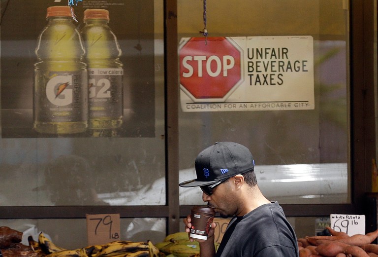 In this July 7, 2014 photo, a man drinks out of a coffee cup as he walks under a sign opposed to taxes on sugary drinks posted in the window of Casa Thai Market in San Francisco. San Francisco and Berkeley are aiming to become the first U.S. cities to pass per-ounce taxes on sugary drinks. (AP Photo/Jeff Chiu)