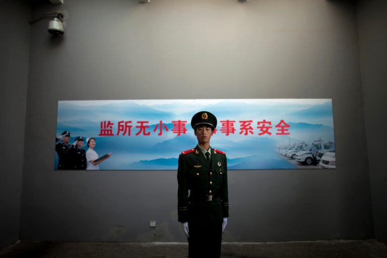 FILE - In this Oct. 25, 2012 file photo, a Chinese paramilitary policeman stands guard underneath a surveillance camera at an entrance to the Number Two Detention Center in Beijing, China. Three police officers and four other people helping them have been convicted of torturing suspects to obtain confessions, China's official state news agency reported, in a rare example of a prosecution of the practice. The slogan on the wall reads 