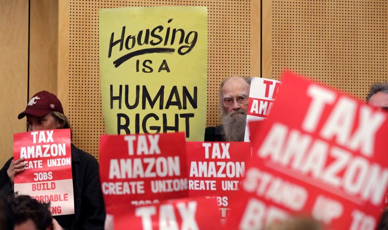 Members of the public look on at a Seattle City Council before the council voted to approve a tax on large businesses such as Amazon and Starbucks to fight homelessness, Monday, May 14, 2018, in Seattle. The council on Monday unanimously backed a compromise tax plan that will charge large businesses about $275 per full-time worker a year. It's lower than the $500-per-worker tax initially proposed. The tax would begin in 2019 and raise about $48 million a year to pay for affordable housing and homeless services. (AP Photo/Elaine Thompson)
