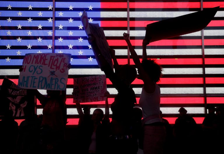 Protesters rally in front of an American flag in Time Square in New York, Thursday, Oct. 4, 2018 over Supreme Court nominee Brett Kavanaugh.