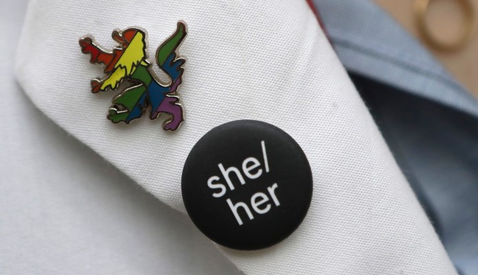 In this Thursday, Oct. 17, 2019 photo Harvard Medical School student Aliya Feroe, of Minneapolis, Minn., displays a button resembling a Harvard School of Medicine coat of arms lion, in rainbow colors that symbolize LGBTQ pride, left, and a button featuring pronouns, center, on the lapel of her lab coat on the schoolâs campus, in Boston.