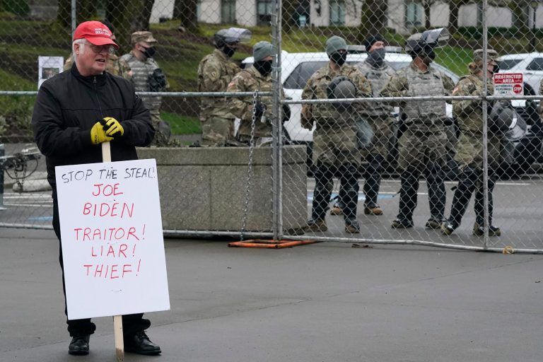 John Hess, of Centralia, Wash., holds a sign opposing President-elect Joe Biden as he stands near Washington National Guard members behind a perimeter fence, Sunday, Jan. 17, 2021, at the Capitol in Olympia, Wash.