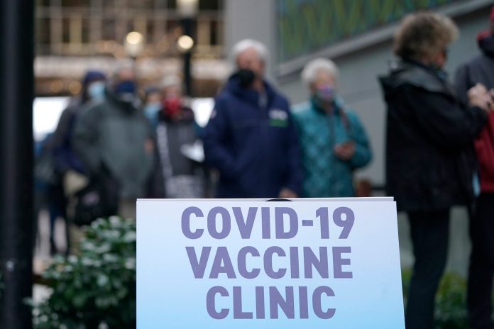 People stand near a sign as they wait in line to receive the first of two doses of the Pfizer vaccine for COVID-19, Sunday, Jan. 24, 2021, at a one-day vaccination clinic set up in an Amazon.com facility in Seattle and operated by Virginia Mason Franciscan Health.