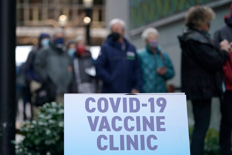 People stand near a sign as they wait in line to receive the first of two doses of the Pfizer vaccine for COVID-19, Sunday, Jan. 24, 2021, at a one-day vaccination clinic set up in an Amazon.com facility in Seattle and operated by Virginia Mason Franciscan Health.