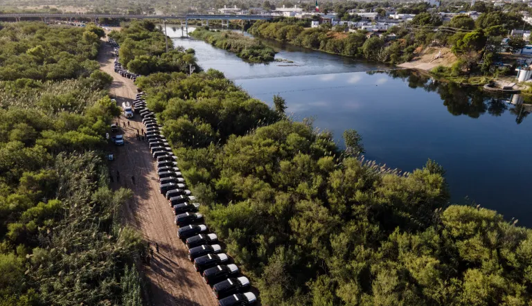A line of Texas Department of Safety vehicles line up on the Texas side of the Rio Grande with Mexico visible, right, near an encampment of migrants.