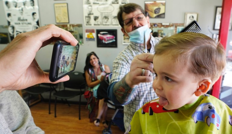 Daniela Garcia, center, observes her son Bobby Miller, 21-months preparing for Motherâs Day by getting his first haircut while watching cartoons on a mobile phone held by his father, Daniel Miller, from barber Mike Douglas at East J Barbers in Sacramento, Calif., Saturday, May 7, 2022.