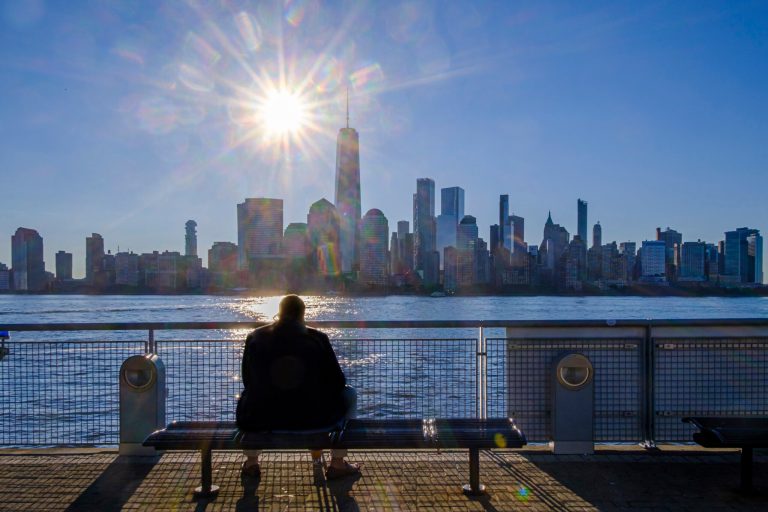 A morning commuter sits on a bench at the ferry terminal in Jersey City, N.J. looking at the lower Manhattan skyline on a cloudless day in New York City, Thursday, Aug. 18, 2022.
