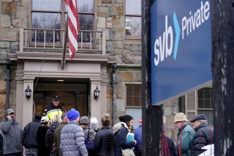 A law enforcement official, behind, stands in an entryway to a branch of Silicon Valley Bank, Monday, March 13, 2023, as customers and bystanders line up outside the branch, in Wellesley, Mass. 