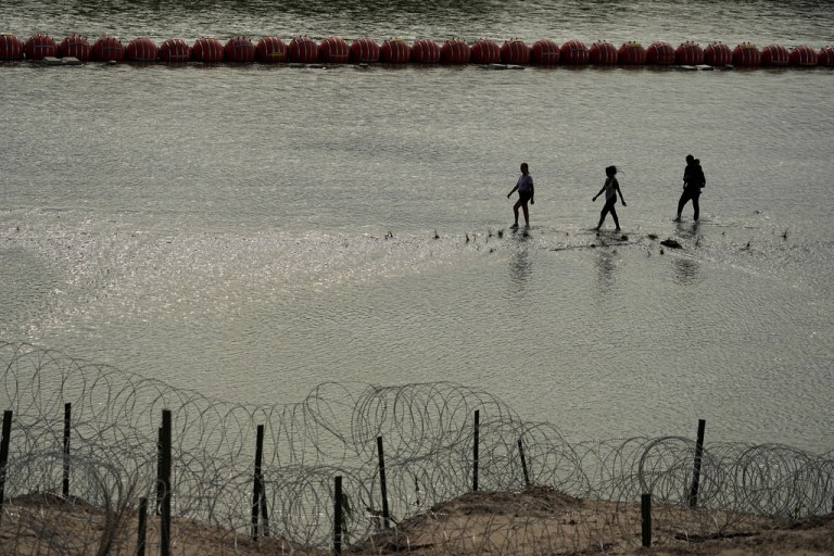Immigrants walk past large buoys being used as a floating border barrier on the Rio Grande.