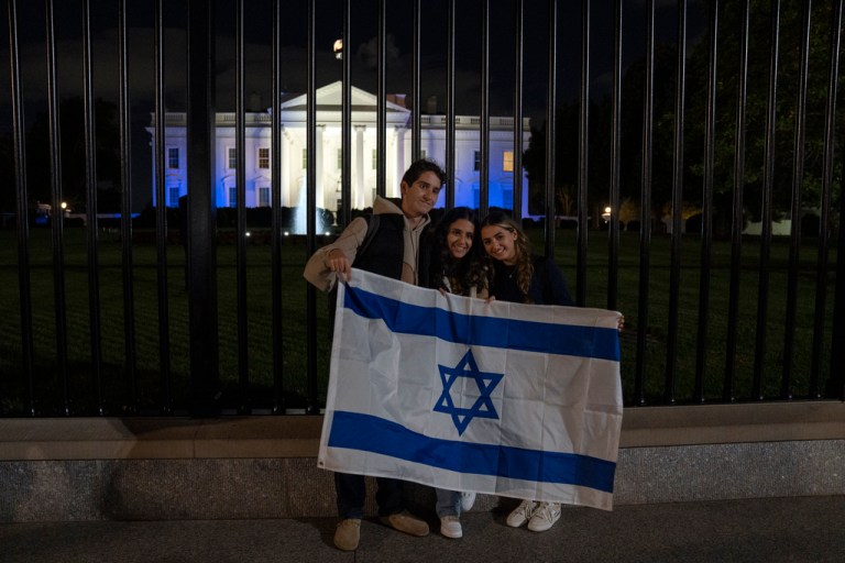 People stand with a Israeli flag outside the White House, lit in blue and white to underscore U.S. solidarity with Israel, Monday, Oct. 9, 2023.