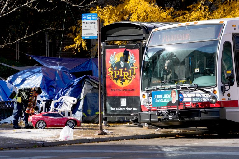 A young migrant boy, left, sweeps off frozen snow on a toy car next to a small tent community and a Chicago Transit Authority bus stop on Wednesday, Nov. 1, 2023, near a Northside police station in Chicago.