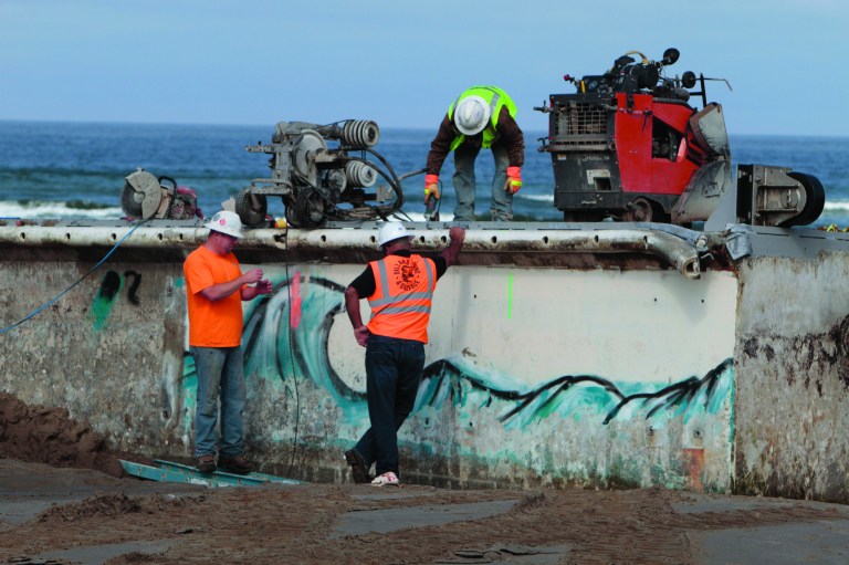 Workers use a cable-cutting system to section the giant Japanese dock that was torn away by last year's tsunami and washed ashore on Wednesday, Aug. 1, 2012 in Newport, Ore. The plan is to cut the 165-ton concrete dock into five slices, like a loaf of bread, and load them onto flatbed trucks, which drive over the soft sand on a roadway of planks and steel plates. (AP Photo/The Oregonian, Randy L. Rasmussen) MAGS OUT; TV OUT; LOCAL TV OUT; LOCAL INTERNET OUT; THE MERCURY OUT; WILLAMETTE WEEK OUT; PAMPLIN MEDIA GROUP OUT