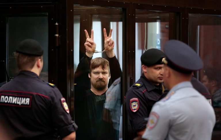 Russian opposition activist Leonid Razvozzhayev, in handcuffs, shows V-signs from a defendant's cage in a court room prior to a hearing in the Bolotnaya square protest trial in Moscow, Russia, Thursday, July 24, 2014.  Sergei Udaltsov and Leonid Razvozzhayev are waiting for the sentence Thursday in the Moscow city court on charges of organizing mass riots and colluding to commit a crime. They both pleaded not guilty in a trial that has dragged on since October 2012, when they were detained and put under house arrest. (AP Photo/Ivan Sekretarev)
