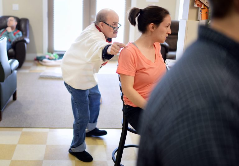 In this photo taken on Wednesday, April 9, 2104, resident Tony Keyt, 52, loves to play pranks on people at the LifeDesigns Highland Group Home in Bloomington, Ind. Here he taps direct support professional Hannah Widmer on the shoulder before sitting down for dinner. Those who care for people with disabilities are frustrated. Indiana Medicaid funding has been cut for the past five years and pay for the state's personal care aides hasn't kept up with inflation. (AP Photo/The Herald-Times, Chris Howell)