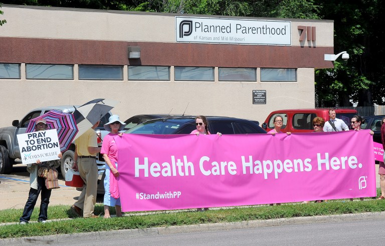 A man holding a sign stands next to five women holding a large banner supporting Planned Parenthood. (Don Shrubshell/Columbia Daily Tribune via AP)
