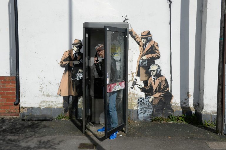 A new graffiti street art piece, suspected of being a Banksy,  appeared in, Cheltenham England  Monday April 14, 2014. Street artist Banksy may have struck again, leaving an espionage-themed graffiti artwork in the hometown of Britain's electronic spy agency.  A stenciled artwork has appeared on a wall in the western England city of Cheltenham, depicting three figures in trench coats and trilbys snooping on a telephone booth.  Cheltenham is home to GCHQ, Britain's electronic eavesdropping agency. The secretive organization found itself in the spotlight after some of its covert activities were revealed by former NSA contractor Edward Snowden. The work has not been claimed on Bansky's official website, but has many of the elusive artist's hallmarks, including the stenciled technique and vein of social commentary. (AP Photo/Ben Birchall/PA) Wire  UNITED KINGDOM OUT  NO SALES  NO ARCHIVE