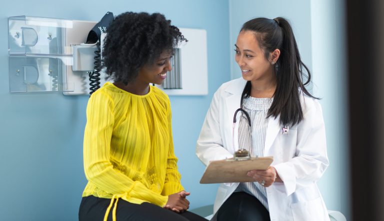 The patient and doctor are sitting next to each other on an examination table in a clinic. The doctor is asking the patient questions and taking notes on a clipboard.