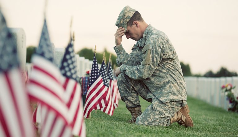 A soldier in ACU fatigues kneels in front of a grave with several American flags in front of it.  