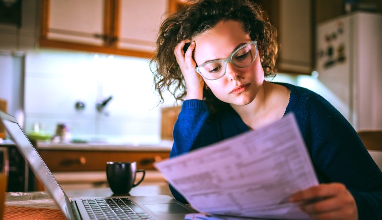 A young woman sadly looks at her bill while sitting in front of a computer.