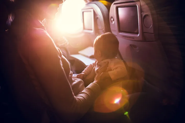 A mother and her baby girl sit in a passenger airline seat, the sun shining brightly in through the plane window. 