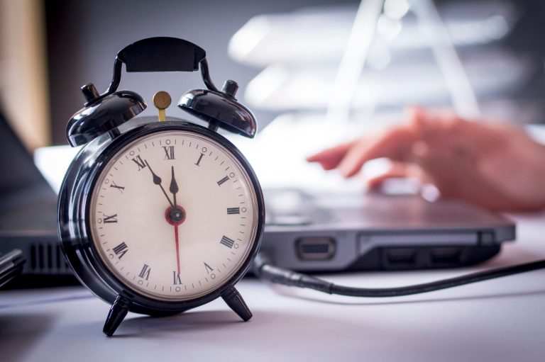 In this stock photo, a person types on a computer, and a large clock with alarm bells sits nearby on the table.