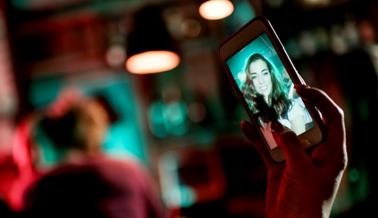 A female hand holds a mobile phone in order to take a selfie image. Other people are in the background of the club.