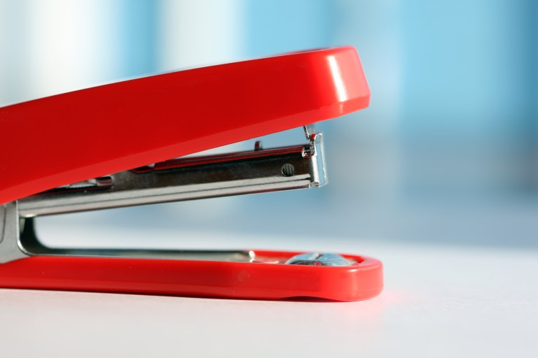A red stapler is pictured sitting on top of a white desk.