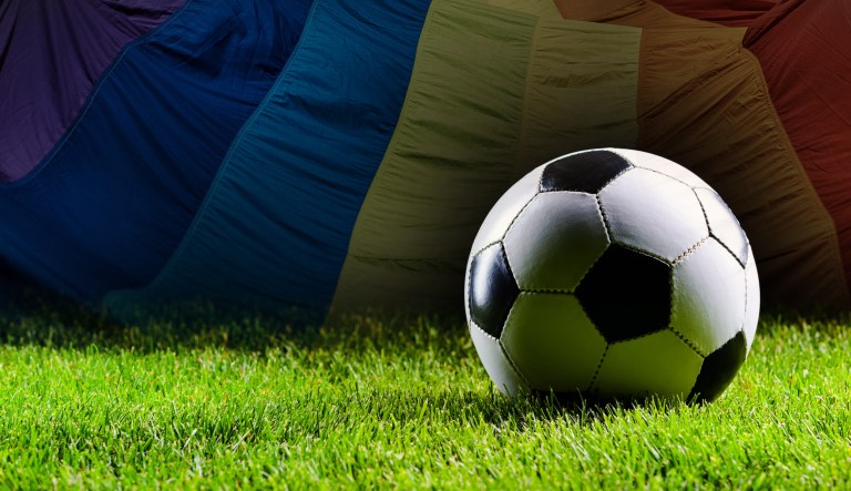 A soccer ball lies on the grass as an LGBT flag is seen in the background.