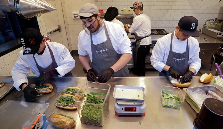 FILE- In this Jan. 22, 2018, file photo workers, as seen from a sidewalk window, make sandwiches in an Amazon Go store in Seattle. Amazon is boosting its minimum wage for all U.S. workers to $15 per hour starting next month. The company said Tuesday, Oct. 2, 2018, that the wage hike will benefit more than 350,000 workers, which includes full-time, part-time, temporary and seasonal positions.