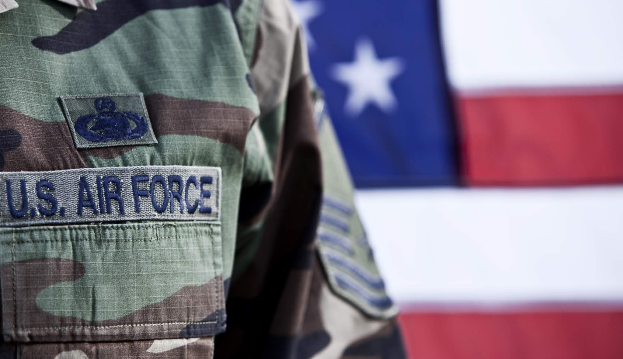 A person is dressed in a U.S. Air Force uniform and stands in front of an American flag.