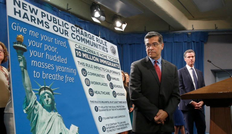 California Attorney General Xavier Becerra, left, glances at a display outlining the supposed effects on immigrants in the state under the Trump administration's new rules blocking green cards for many of those who receive government assistance, during a news conference in Sacramento, Calif., Friday, Aug. 16, 2019.