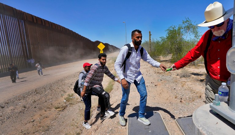 Retired schoolteacher Tom Wingo of Samaritans Without Borders, right, gives snacks and bottles of water to a group of migrants claiming to be from India, who just crossed the border wall, Tuesday, Aug. 29, 2023, in Organ Pipe Cactus National Monument near Lukeville, Arizona. 