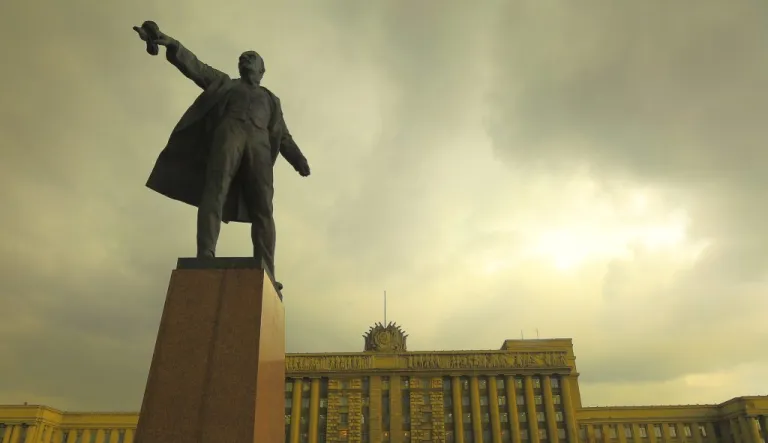 A statue stands tall in front of a building with the man's hand out in the air against a cloudy gray-yellow sky.