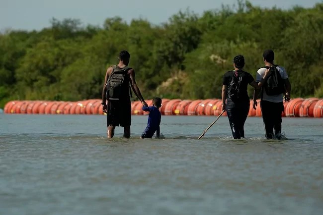 Migrants crossing into the U.S. from Mexico walk along large buoys being used as a floating border barrier on the Rio Grande Tuesday, Aug. 1, 2023, in Eagle Pass, Texas.