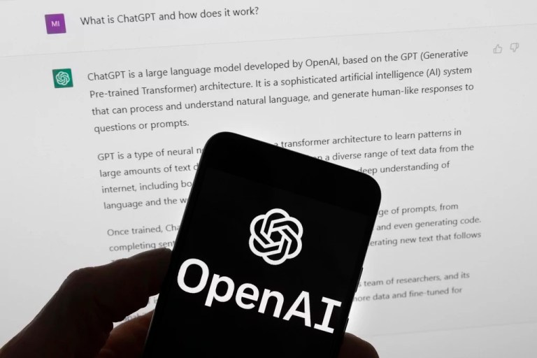 The OpenAI logo is seen on a mobile phone in front of a computer screen which displays output from ChatGPT.