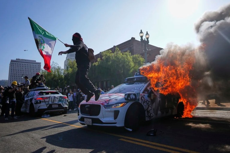 A protester leaps off a burning Waymo taxi near the Metropolitan Detention Center in downtown Los Angeles