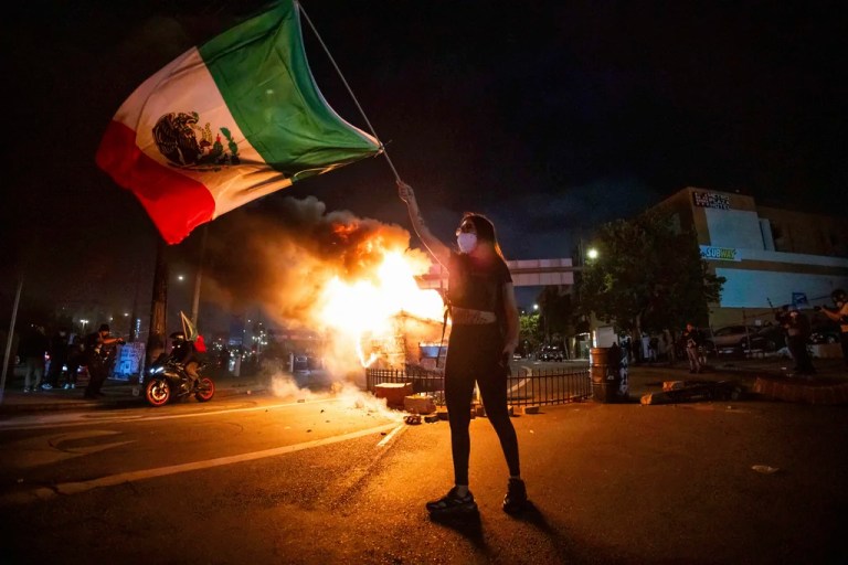 A woman waves the Mexican flag as flames erupt from a burning dumpster during a protest in downtown Los Angeles, Sunday, June 8, 2025.