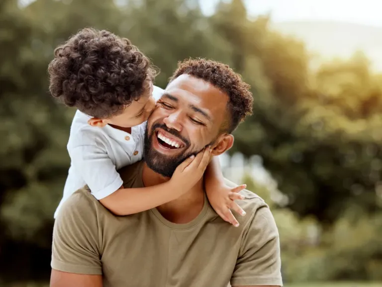 A father smiles as his son hugs him around his neck and kisses his cheek.