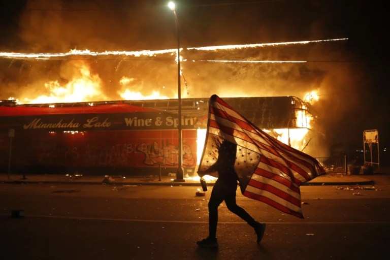 In this May 28, 2020, file photo, a protester carries an American flag upside down, a sign of distress, next to a burning building in Minneapolis.