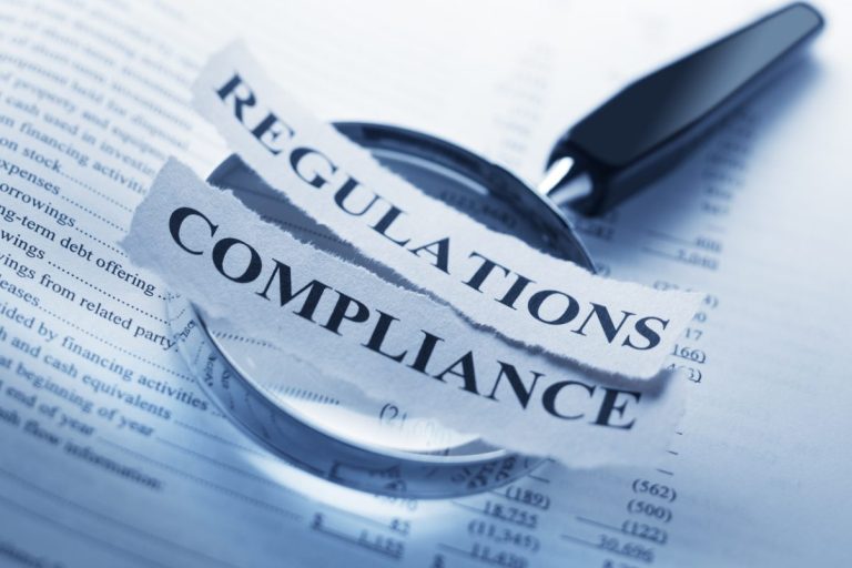 The words Regulation and Compliance are printed on a torn piece of paper that sits on top of a magnifying glass which sits on a corporate financial document. The image illustrates the concept of government oversight and corporate and financial governance.