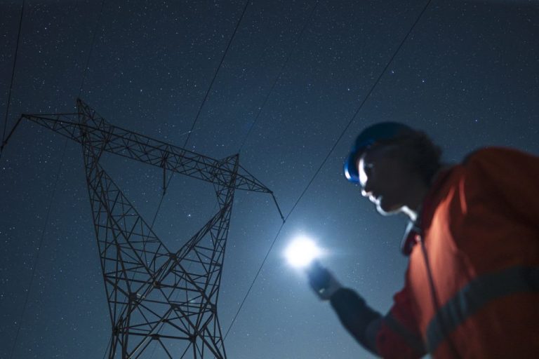 Electrical engineer with safety jacket and helmet inspects high voltage electric pylons, checking the condition of electrical components at night using flashlight.