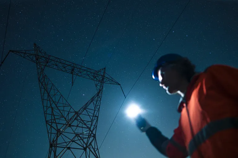 Electrical engineer with safety jacket & helmet inspects high voltage electric pylons, checking the condition of electrical components at night using flashlight
