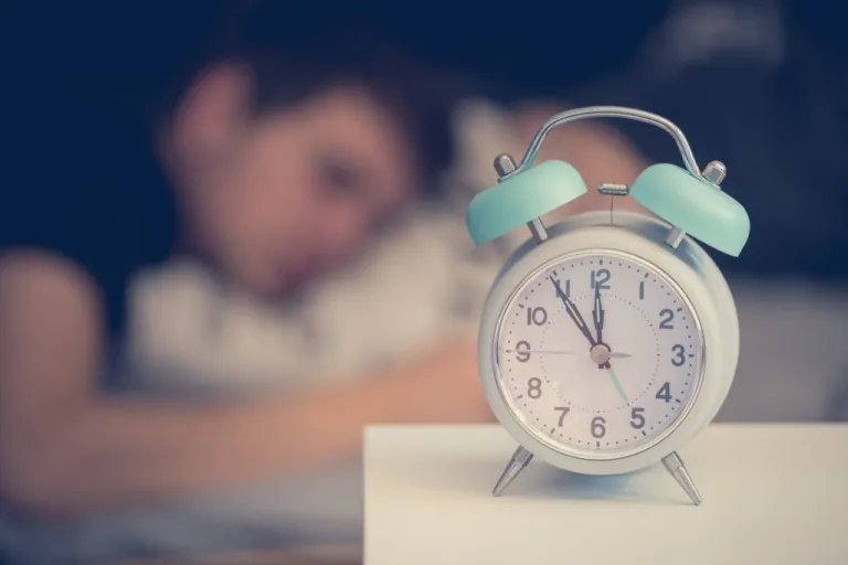 White alarm clock on a bedside table with a young man sleeping in the background.