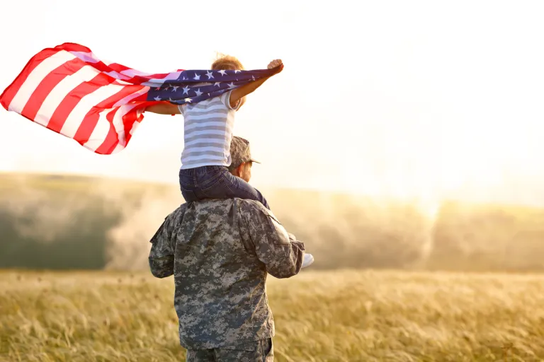 A military man carries his son, who is holding an American flag, on his shoulders in a sunny field.