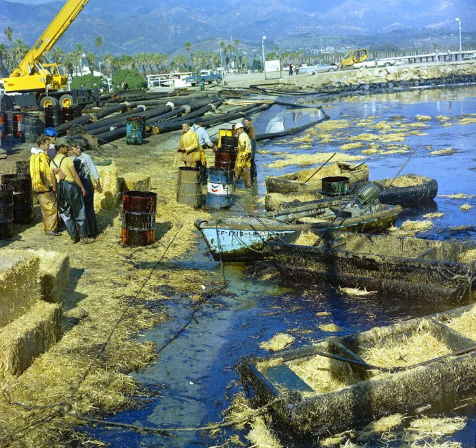 Workers using pitchforks, rakes, and shovels attempt to clean up oil-soaked straw from a beach.