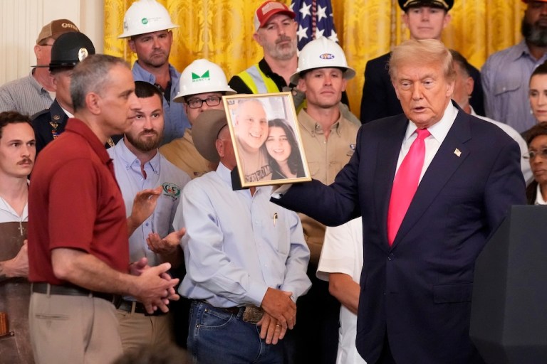President Donald Trump holds a photo of Katie Abraham, pictured right, as her father Joe Abraham, left, watches during an event to promote Trump's domestic policy and budget agenda in the East Room of the White House.