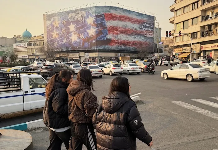 People walk in front of a billboard with a graphic showing a U.S aircraft carrier with damaged fighter jets on its deck, and sign reading in Farsi and English: 