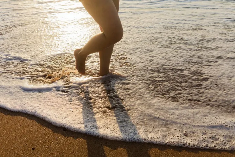 A person walks barefoot into the calm, shallow waves of the ocean at sunset.