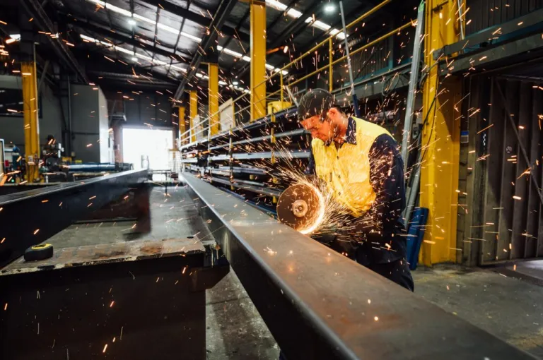 Industrial worker in a workshop wearing safety gear using a grinder emitting sparkse in large industrial warehouse with machinery and yellow support structures under bright overhead lighting.