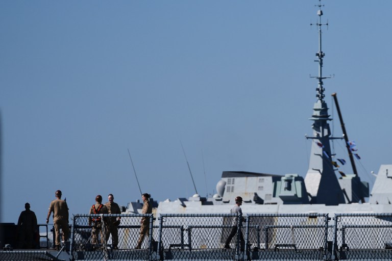 A French frigate enters the port behind sailors walking.